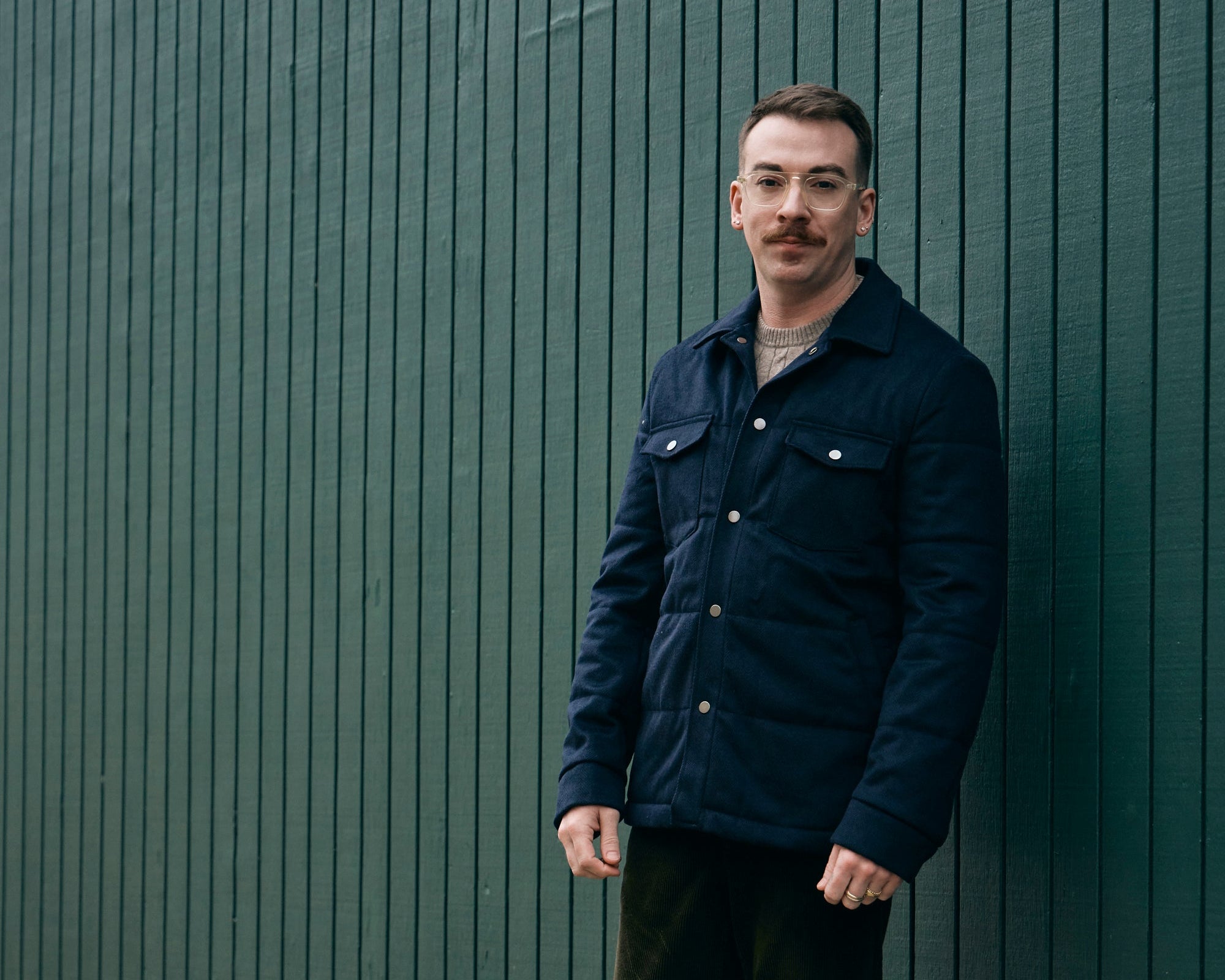 Man wearing a navy jacket standing against a green wall.